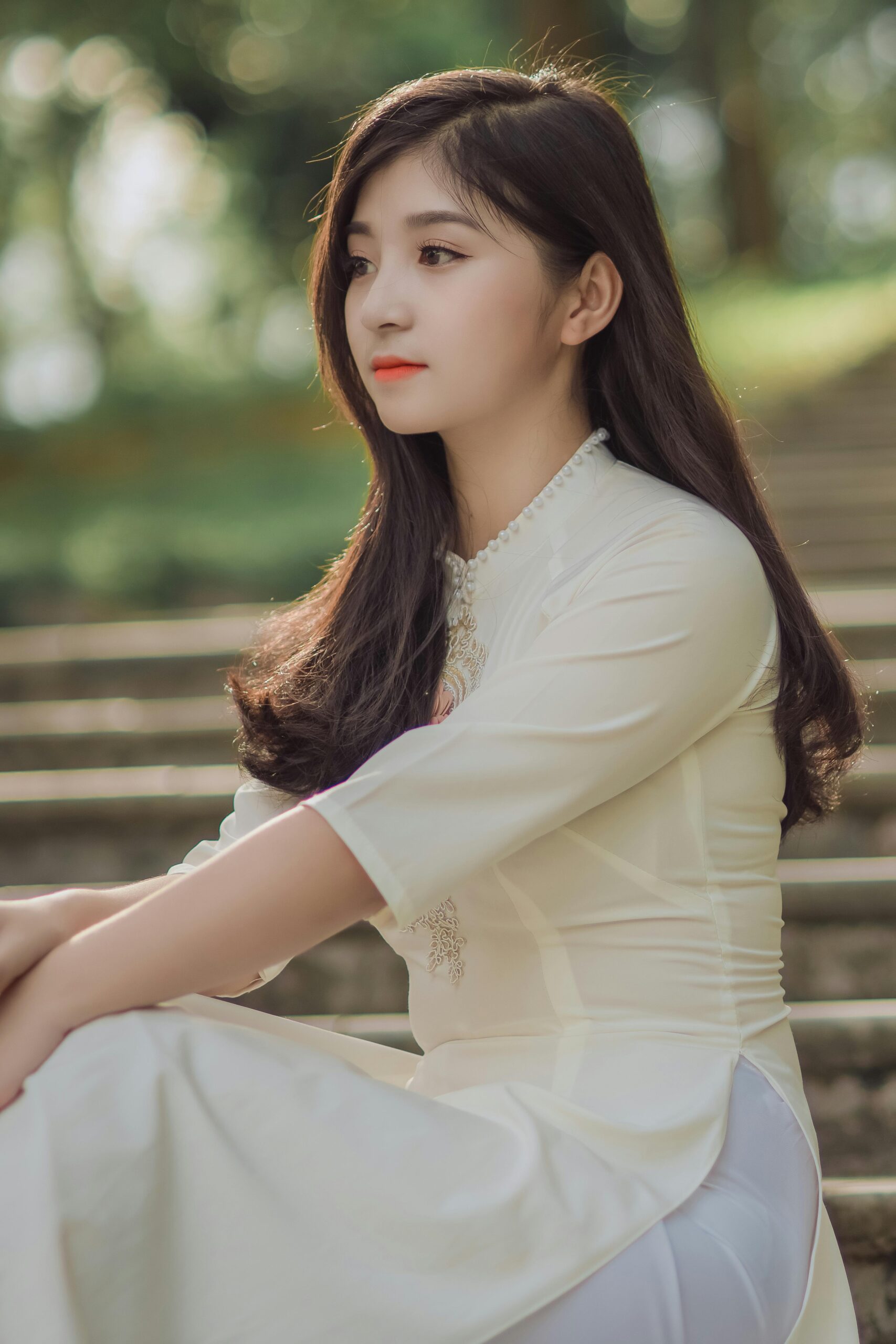 Asian woman in a white dress sitting on steps outdoors, showcasing elegance and beauty.