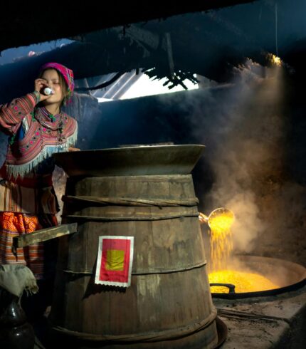 Woman in traditional ethnic attire cooking food in a rural setting with a large cauldron.
