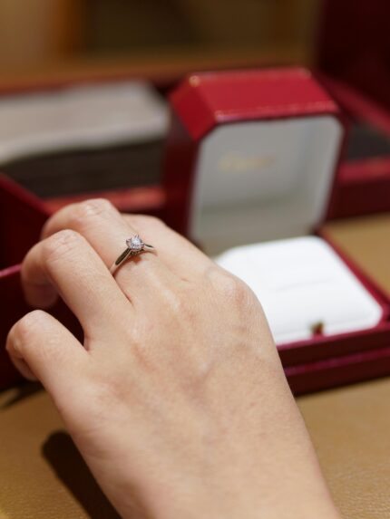 Close-up of a hand with an elegant diamond engagement ring against a blurred jewelry box background.