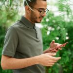 Young man checks phone surrounded by lush green plants, evoking a natural setting.