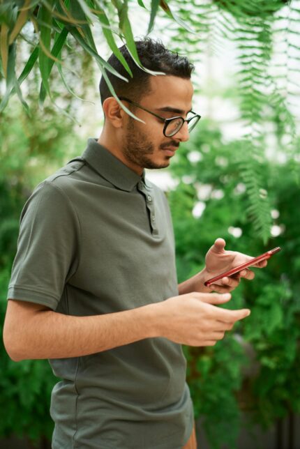 Young man checks phone surrounded by lush green plants, evoking a natural setting.