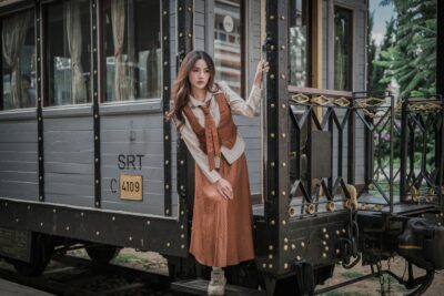 Elegant woman in vintage attire posing by a classic train carriage gently holding the handle.