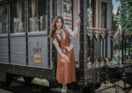 Elegant woman in vintage attire posing by a classic train carriage gently holding the handle.
