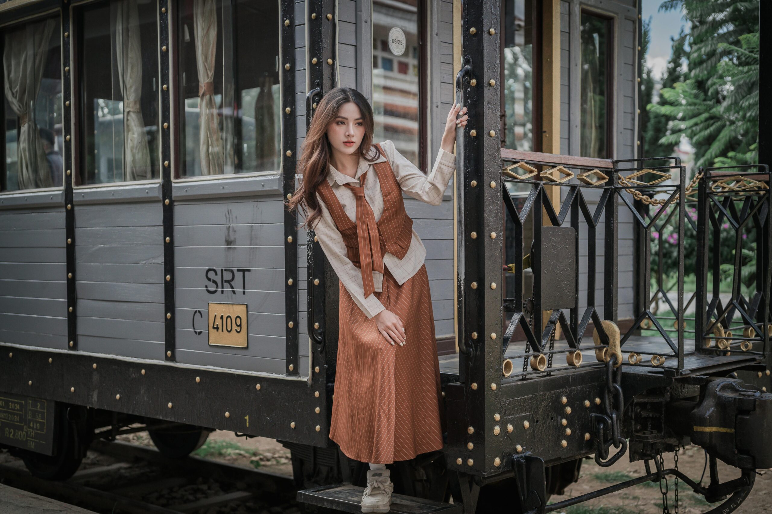 Elegant woman in vintage attire posing by a classic train carriage gently holding the handle.