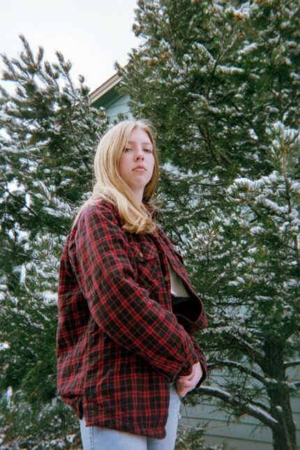 Teenager in plaid jacket standing outdoors among snow-covered trees, winter setting.