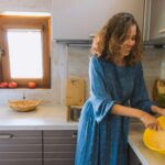Smiling woman in blue dress mixes dough in cozy kitchen with sunlight streaming through window.