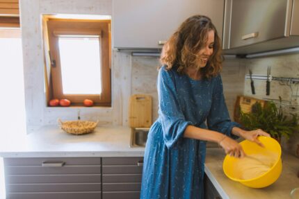Smiling woman in blue dress mixes dough in cozy kitchen with sunlight streaming through window.