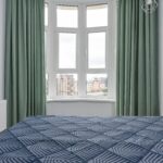Interior of cozy light bedroom with blue blanket on bed placed against window with green curtain and white walls at home