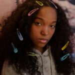 Portrait of a teenager with curly hair adorned with colorful hair clips, wearing a grey hoodie.