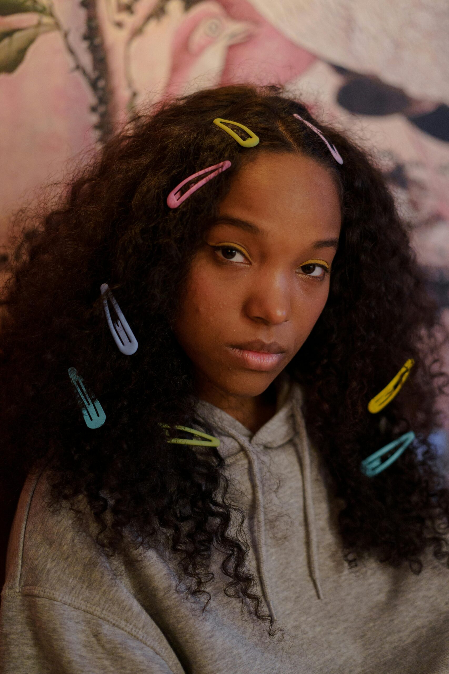Portrait of a teenager with curly hair adorned with colorful hair clips, wearing a grey hoodie.