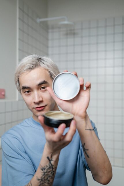 A young man with blonde hair shows off a hairstyling product in a modern bathroom.