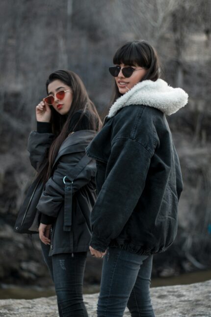 Two stylish women in denim jackets and sunglasses pose outdoors, showcasing winter fashion.