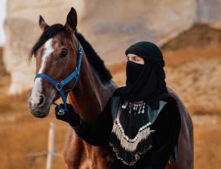 A woman in traditional dress with a horse in a desert landscape.
