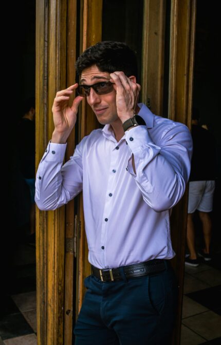 Confident man in sunglasses and smart attire adjusting glasses by a wooden doorway.