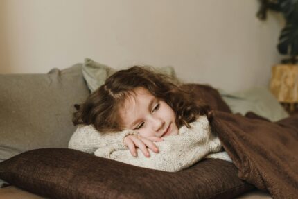 A child in a knitted sweater rests comfortably on a couch with pillows and a blanket.