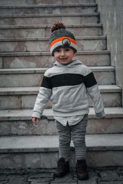 Adorable toddler in a knit hat smiling on stone steps, wearing a gray hoodie and leggings.