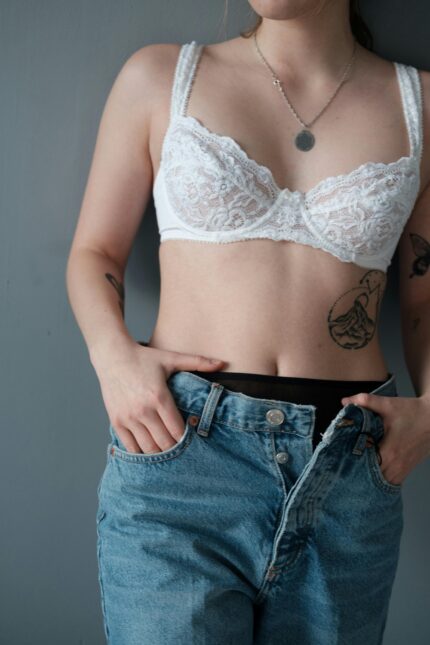 Unrecognizable slim woman with tattoo wearing white lingerie and unbuttoned denim shorts standing on gray background with hands in pockets in studio